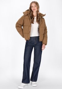 Brun puffjakke med en aftagelig hætte, der har quiltede paneler og trykknapper, kombineret med højtaljede mørkeblå jeans og hvide sneakers.