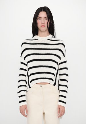 Maglione a righe bianche e nere in maglia, con scollatura a girocollo e design corto. Il modello lo abbina a pantaloni beige a vita alta.