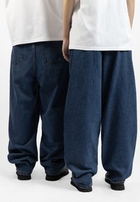Avslappede, mørkeblå denimbukser med tapered ben, synlige baklommer og en løs, slouchy silhuett. Båret med svarte sko.