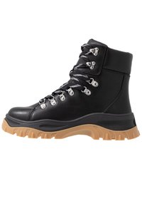 Bota de senderismo de piel negra con tobillo alto, suela de goma texturizada, herrajes plateados y suela de goma en contraste en color marrón; presenta cordones en blanco y negro.