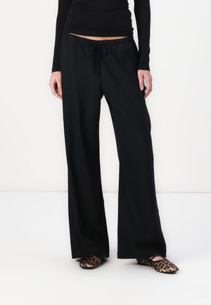 Model trägt lockere schwarze Hose mit Kordelzug, kombiniert mit flachen Schuhen im Leopardenmuster, stehend vor einem einfarbigen weißen Hintergrund.