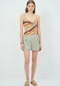 Brun og gulstribet camisole-top med draperet halsudskæring, parret med lysegrå, plisserede shorts. Modellen står på en neutral baggrund.
