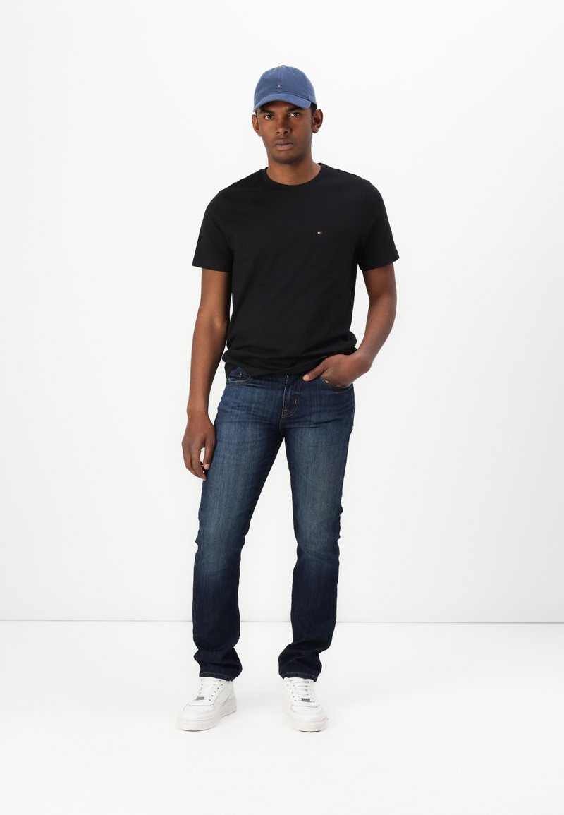 Czarny t-shirt z okrągłym dekoltem, niebieska czapka, ciemnoniebieskie dżinsy i białe trampki. Luźny strój z widocznym logo na koszulce.