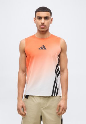 Uomo che indossa una canotta sportiva sfumata dall'arancione al bianco con strisce nere e logo, pantaloncini beige, in piedi davanti a uno sfondo uniforme.