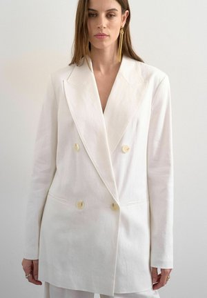 Donna che indossa un blazer doppiopetto bianco con ampi rever e orecchini a frangia con catene dorate, su uno sfondo chiaro e semplice.