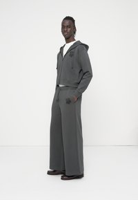 Γκρι κοντοκουρεμένο φούτερ με λογότυπο λουλουδ ιού, σε συνδυασμό με γκρι παντελόνια φαρδιάς γραμμής. Το υλικό φαίνεται μαλακό. Το μοντέλο στέκεται σε απλό λευκό φόντο.