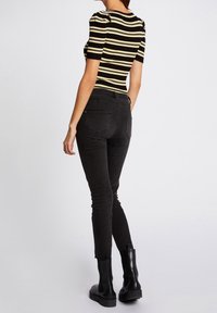 Eine Frau trägt ein kurzärmliges Oberteil mit schwarzen und beigen Streifen, eng anliegende schwarze Jeans und schwarze Stiefeletten. Sie steht vor einem einfarbigen Hintergrund.
