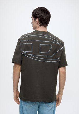 Homme portant un t-shirt vert foncé avec un grand motif abstrait bleu clair dans le dos, accompagné d’un jean bleu clair, de dos.