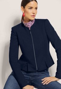 Marineblaue Peplum-Jacke mit einem Frontreißverschluss, ausgestattet mit detaillierten Nähten und einem hohen Kragen, kombiniert mit einem gemusterten Hemd darunter.