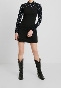 Abito nero senza maniche con scollo quadrato sopra una maglietta a maniche lunghe a fantasia; abbinato a stivali da cowboy neri con punta appuntita e cuciture decorative.