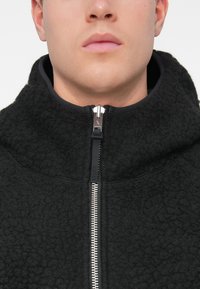 Casaco de polar preto com gola alta, fecho éclair prateado, tecido texturado e forro preto em contraste ao longo da área do zíper.