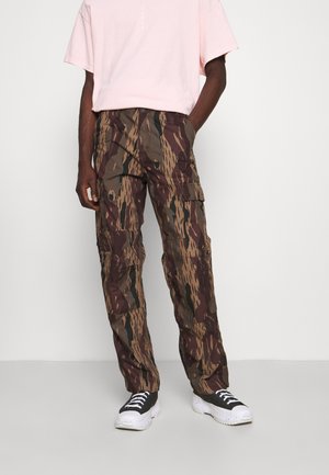 Osoba ubrana w brązowo-zielone moro spodnie typu cargo, czarno-białe trampki oraz jasnoróżową koszulkę z krótkim rękawem, stojąca na jednolitym tle.