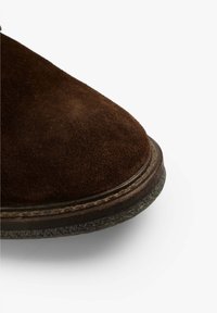 Zapato de ante marrón, punta redonda, superficie texturizada, detalle cosido a lo largo de la suela, suela de goma oscura que contrasta.