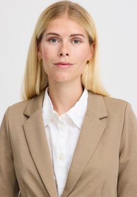 Beež bleiser, millel on sisselõigatud krae, valge nööbitava särk üleval. Kangas näib siledana, hästi istuvas lõikes. Lihtne, elegantne disain.