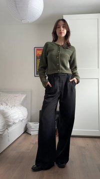 Mujer joven de pie en un dormitorio, vistiendo un cárdigan verde, pantalones anchos negros y zapatos negros, con las manos en los bolsillos.