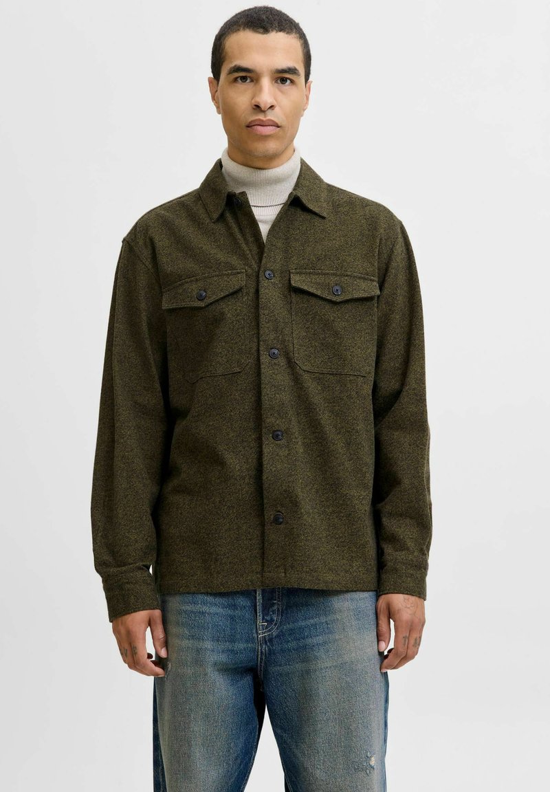 Mand iført en olivengrøn knap-vest over en beige turtleneck og falmede blå jeans, stående mod en hvid baggrund.