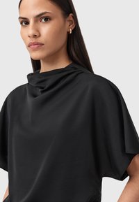 Top nero drapeggiato con collo alto, texture morbida e vestibilità ampia. Presenta dettagli laterali e un design minimalista.