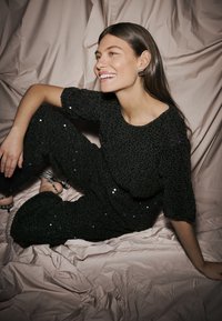 Outfit in maglia nera decorato con paillettes, maniche corte e pantaloni larghi, abbinato a orecchini a cerchio argentati, su uno sfondo rosa chiaro.