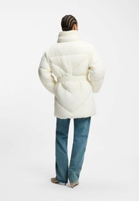 Vit pufferjacka med hög krage, kviltat mönster, bälte i midjan och oversized passform. Tillsammans med ljusblå jeans och beige skor.