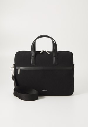 Sort taske med et håndtag på toppen, lavet af lærredsmateriale, med tekstureret overflade, sølvfarvede lynlåse, aftagelig skulderrem og et Calvin Klein-logo i bunden.