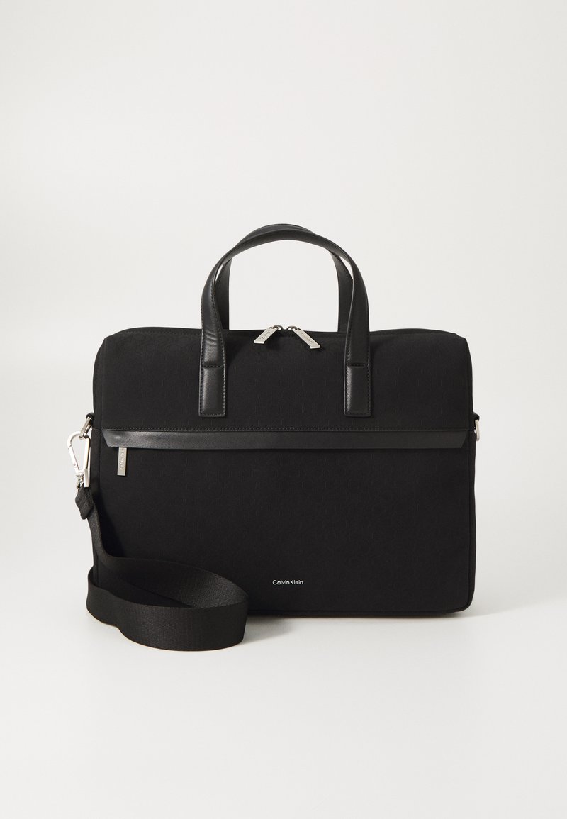Black bag with a top handle, canvas material, textured surface, silver zippers, detachable shoulder strap, and a Calvin Klein logo at the base.