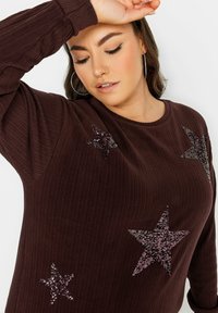 Brauner Rippstrickpullover mit dekorativen, glitzernden Sternen in Dunkel- und Helllila. Das Design umfasst einen runden Halsausschnitt und lange Ärmel.
