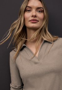 Jeune femme aux longs cheveux blonds portant un pull beige côtelé à col en V, debout devant un fond gris foncé.