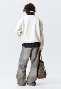 Chaqueta de forro polar blanca con cuello negro, combinada con pantalones vaqueros grises holgados. Llevando una bolsa de tote marrón con un pañuelo a cuadros.