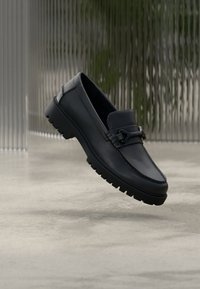 Παπούτσι από σκούρο μπλε δέρμα χωρίς κορδόνια, με υφασμένη λωρίδα και ασημί διακοσμητικά. Διαθέτει παχιά, ανθεκτική μαύρη λαστιχένια σόλα. Επιπλέει πάνω σε ουδέτερο φόντο.