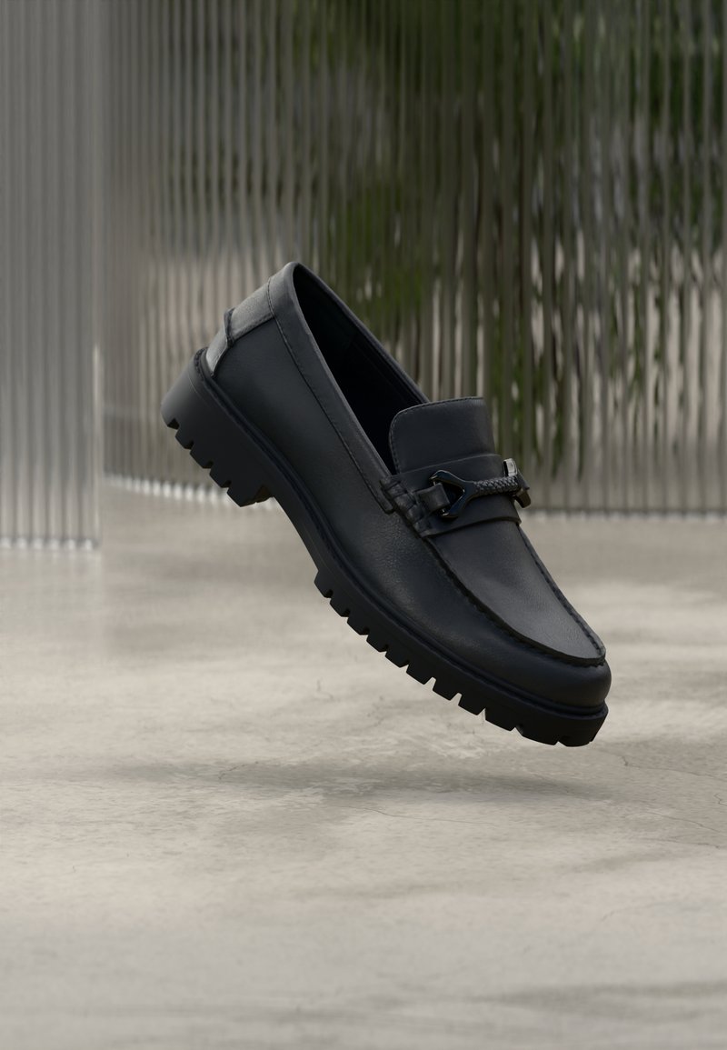 Mocassin en cuir bleu marine avec une bride texturée et des accents métalliques argentés. Dispose d'une semelle épaisse et robuste en caoutchouc noir. Flotte sur un fond neutre.