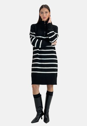 Kvinde står med hånden på ansigtet iført sort og hvidt vandret stribet sweaterkjole og knæhøje sorte støvler med hæl.