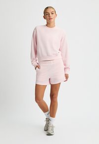 Felpa rosa chiaro con collo a giro e polsini a coste, abbinata a pantaloni corti coordinati. Entrambi presentano una texture morbida e liscia.
