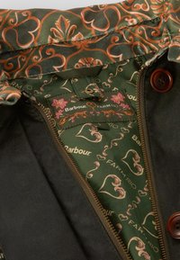 Interior de uma jaqueta castanha e verde com padrões florais e de corações, apresentando um fecho de correr dourado e botões castanhos. Etiquetas visíveis: "Barbour" e "FARMRIO."
