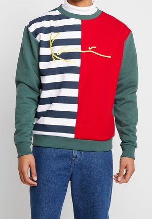 Homme portant un sweat-shirt avec des manches vertes, des rayures horizontales bleu marine et blanches d’un côté, rouge uni de l’autre, et un lettrage jaune sur la poitrine.