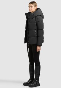 Femme debout portant une doudoune noire à capuche, des leggings noirs et des bottes noires montant jusqu'aux genoux, sur un fond clair uni.