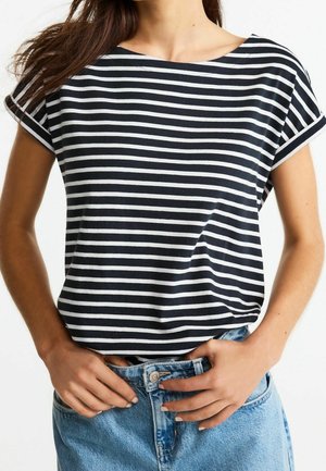 Vrouw draagt een zwart-wit gestreept T-shirt met korte mouwen, horizontale strepen, ingestopt in een hoge taille lichtblauwe spijkerbroek, met handen dichtbij de tailleband.