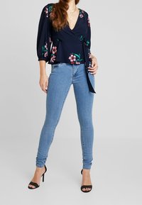 Blusa navy con estampado floral y cintura atada, combinada con jeans skinny de color azul claro y sandalias de tacón negras. Tejido suave, diseño ajustado.