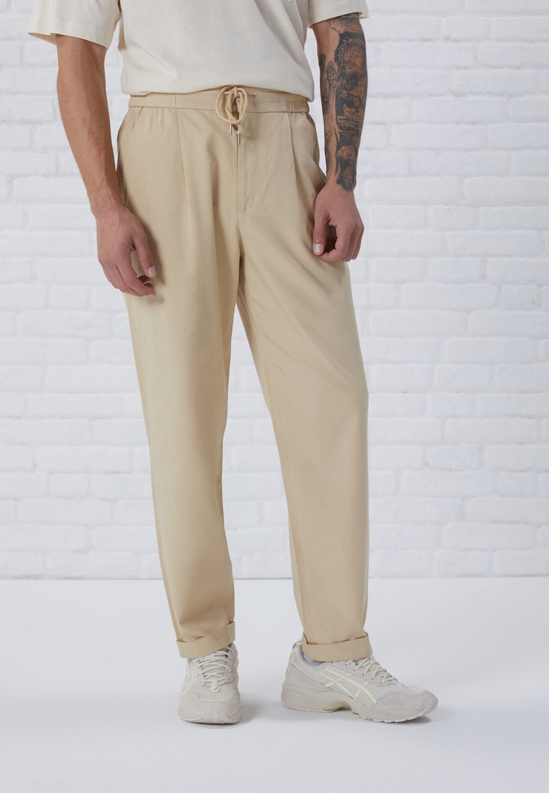 Pantalons beige avec une coupe décontractée, taille élastique à coulisse, plis et ourlets roulés, associés à des chaussures de sport gris clair.
