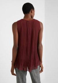 Burgundy ärmlös plisserad topp med rund halsringning, en nyckelhålsknappar på baksidan och en flödande nederkant. Mjuk, lättvikts tyg.