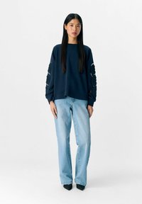 Marineblauwe sweatshirt met ronde, gestructureerde patronen op de mouwen, gecombineerd met lichtblauwe jeans en zwarte puntige hakken. Gladde stof, ontspannen pasvorm.
