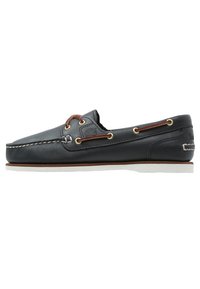 Scarpa da barca in pelle nera con suola in gomma bianca, dettagli marroni, occhielli circolari e cuciture a contrasto. Presenta un logo sul lato.