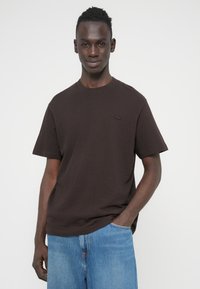 Καφέ βαμβακερό t-shirt με άνετη εφαρμογή, στρογγυλή λαιμόκοψη και κοντά μανίκια. Διαθέτει μικρό, τονικό λογότυπο στο στήθος. Καθημερινό σχέδιο.