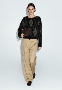 Maglione lavorato a maglia argyle in nero e grigio, pantaloni beige a gamba larga e pochette marrone scuro. Texture lisce e vestibilità rilassata.