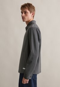 Szara marynarka w kratę z usztywnionym kołnierzem, długimi rękawami, przednią kieszenią i guzikami przy mankietach. Noszona na niebieskich spodniach.