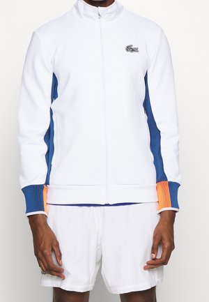 Męska biała sportowa kurtka na zamek z niebiesko-pomarańczowymi prążkowanymi panelami bocznymi i mankietami, zestawiona z białymi krótkimi spodniami sportowymi.