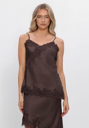 Femme portant un caraco en satin brun foncé et une jupe assortie avec bordure en dentelle, debout devant un fond clair uni.