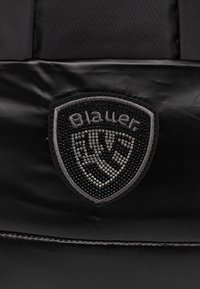 Černá textilní nášivka s vyvýšeným nápisem "Blauer" a krystalovými akcenty, umístěná na hladkém černém pozadí.