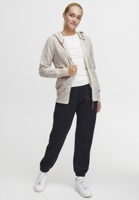 Hettegenser i lys beige med fargerike prikker, sammen med mørke joggebukser. Har glidelås og ribbestrikkede mansjetter og liv.