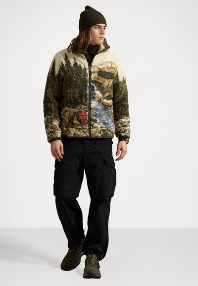 Fleece jacket with nature scene design, featuring trees, a river, and rocks. Dark green with a buttoned pocket, worn with black cargo pants.