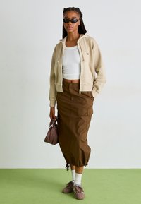 Béžová mikina na zips, biely rebrovaný crop top, hnedá cargo sukňa so sťahovacím lemom a hnedé tenisky. Doplnok: malá hnedá taška.